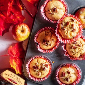 Maple Glazed Apple Cider Pecan Muffins in the muffin tin with fall leaves apples and cinnamon stick beside