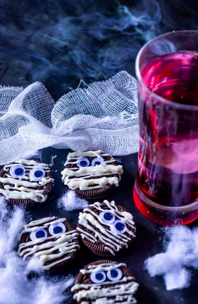 Halloween Mummy Oreos arranged on a black background with a glass of red drink beside it and smoke in the background