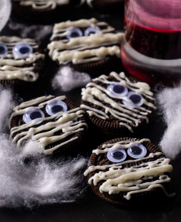 Five Halloween Mummy Oreos on a table with a little cotton spread beside it and a glass of red drink whose base is visible kept beside it