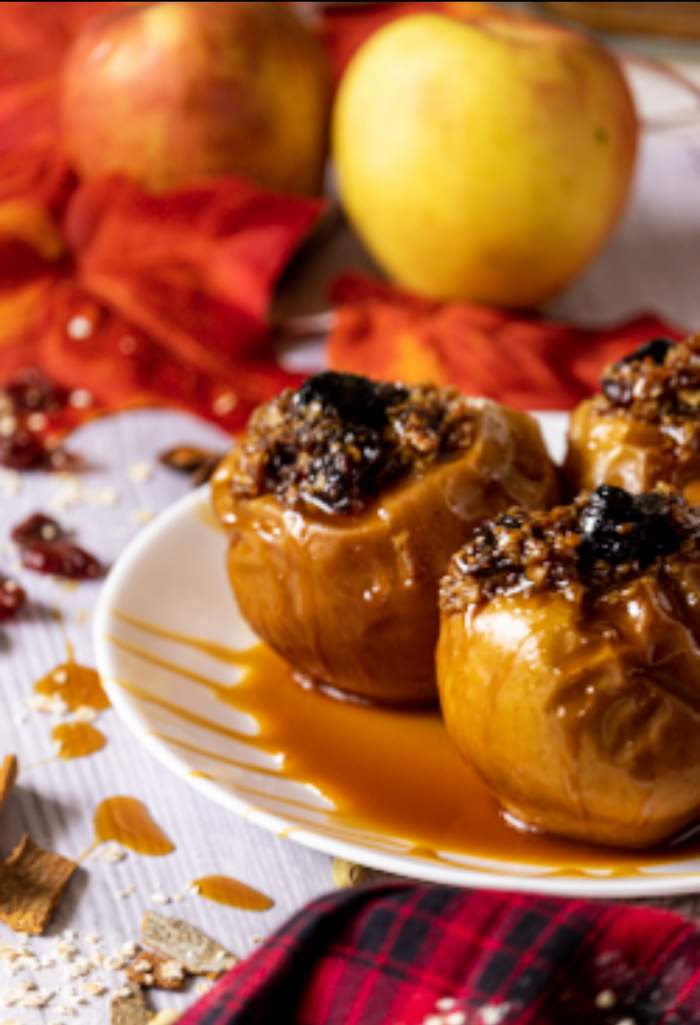 Baked apples on a white plate with fall leaves and fresh apples in the background and a red plaid cloth in the front